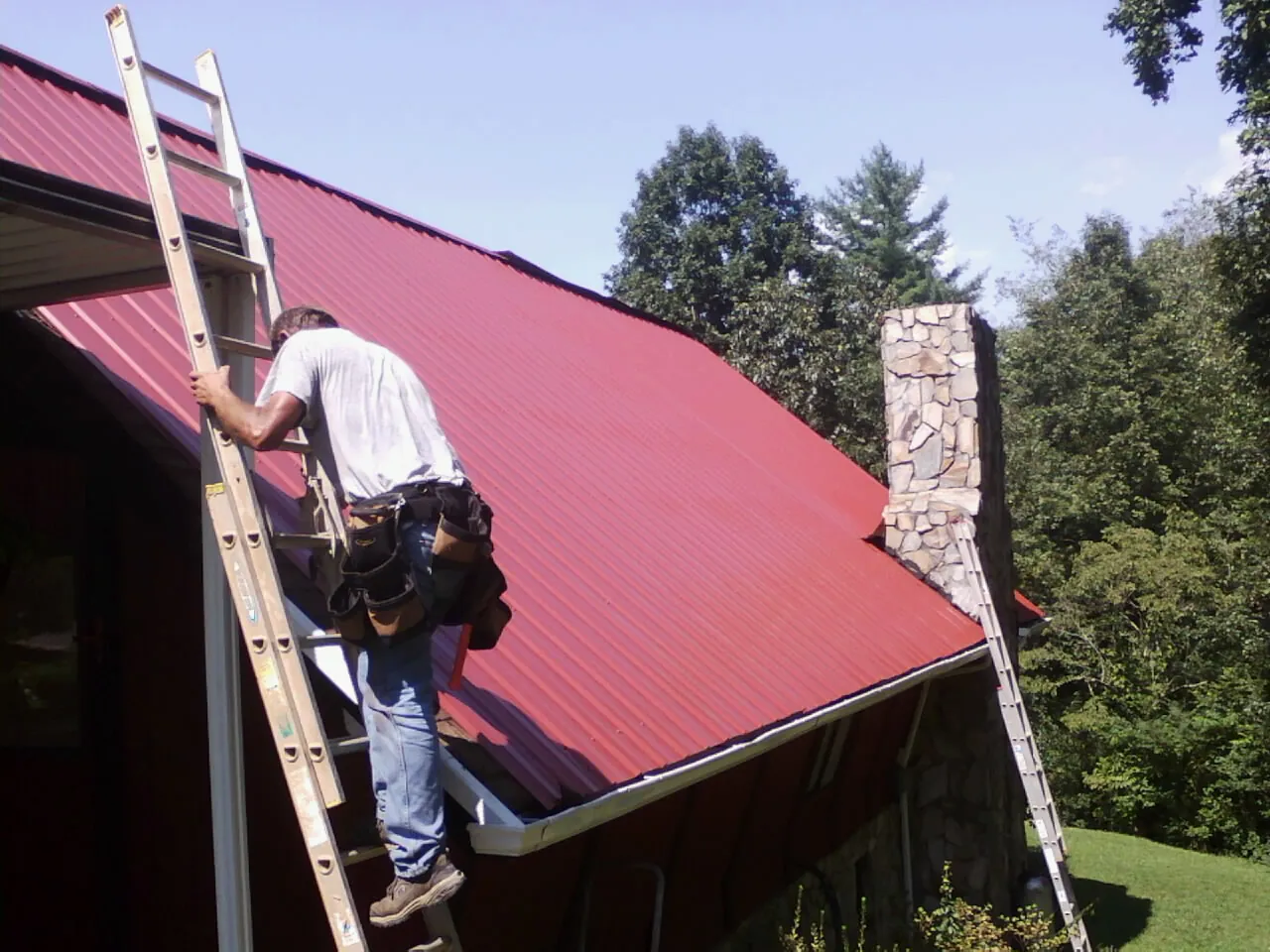 Roofer on a ladder beside a red metal roof and stone chimney — Soffit & Fascia Repair by Roofing Brothers in Groveport